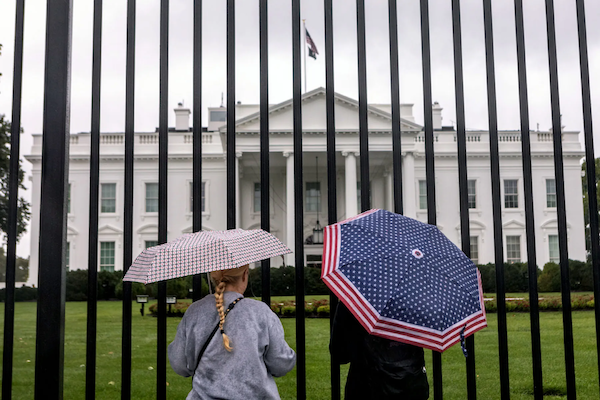 Two people with umbrellas outside the White House, viewed through iron bars.