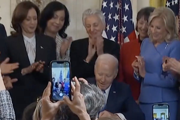 A group of people in a room with American flags, some recording with their phones.