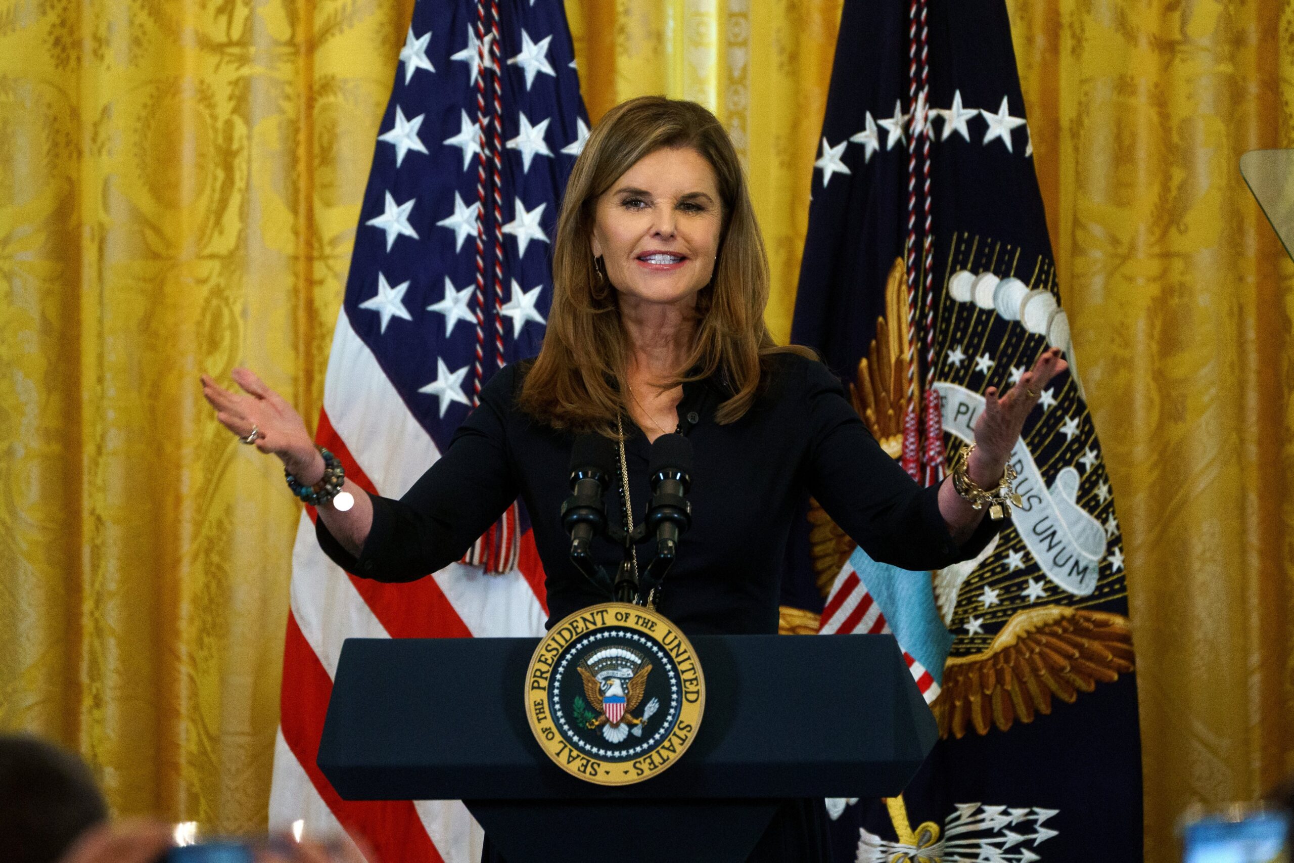 Maria Shriver at the podium with the Presidential Seal, US flags and a gold curtain behind her.