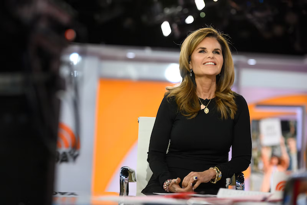 Maria Shriver seated at a desk with a microphone, wearing a black top and accessories.