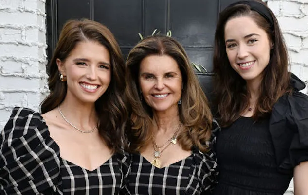 Maria Shriver and her two daughters smile and pose together in front of a white brick wall.