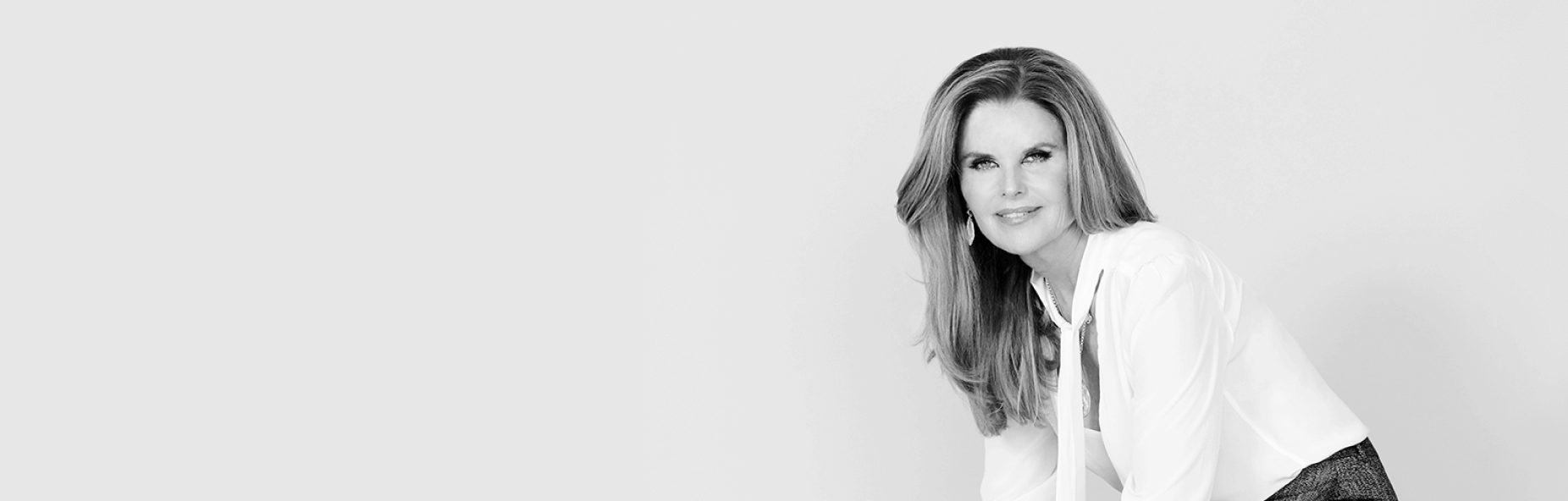 Maria Shriver in white shirt and gray pants seated on a cube, in a grayscale photo.