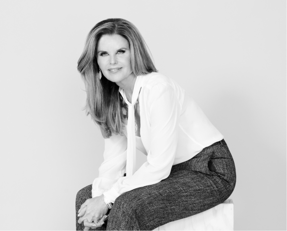 Maria Shriver in white shirt and gray pants seated on a cube, in a grayscale photo.