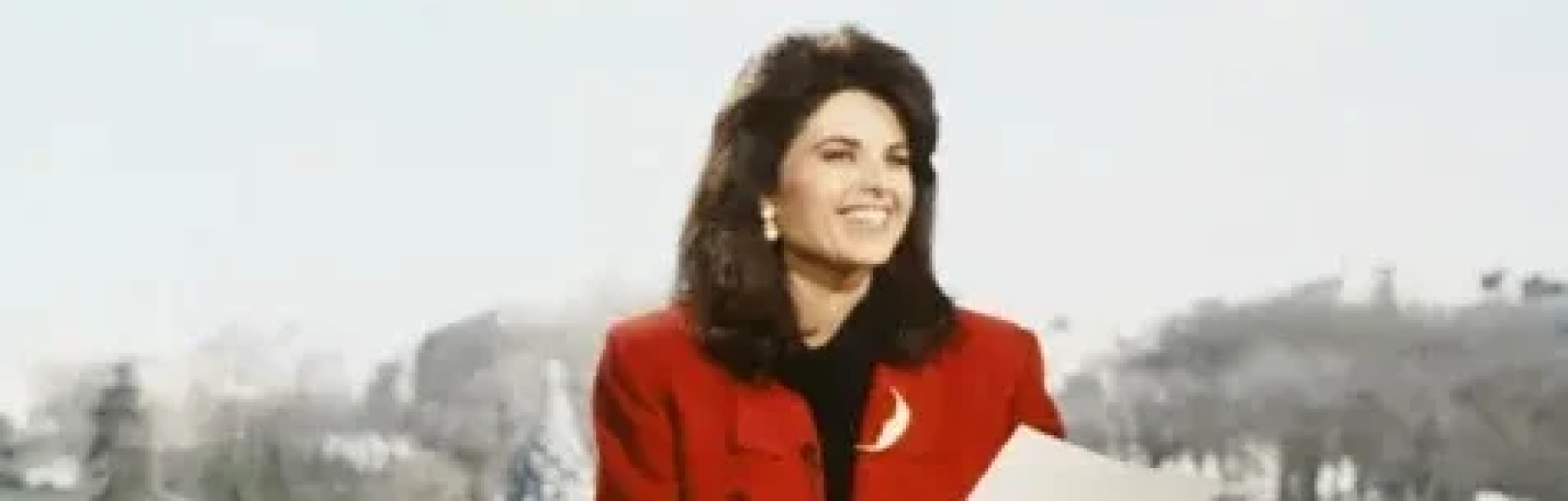 Maria Shriver in red coat reading a paper with a blurred background.