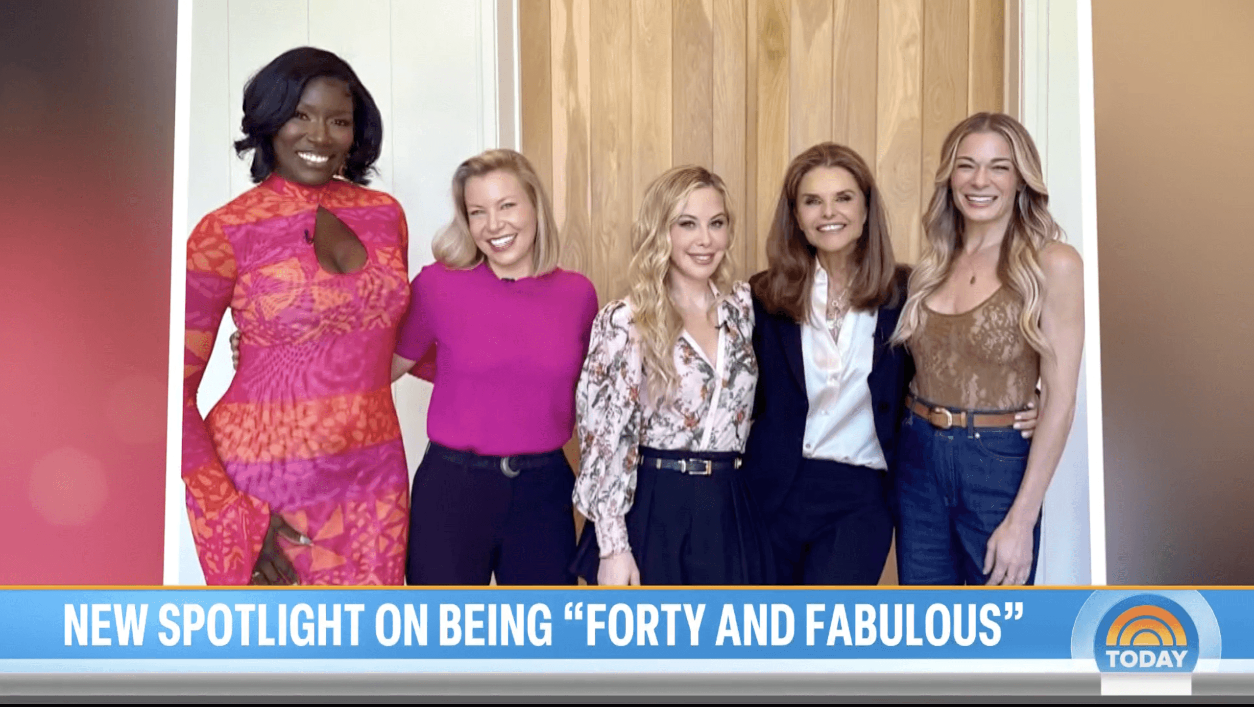 Five women stand side by side smiling for a group photo in front of a wooden door. From left to right: a woman in a bright pink and orange patterned dress, a woman in a hot pink top and dark pants, a woman in a floral blouse with a navy skirt, a woman in a navy blazer over a white blouse, and a woman in a tan sleeveless top with blue jeans. A banner at the bottom reads, “New Spotlight on Being ‘Forty and Fabulous,’” with the TODAY show logo in the lower-right corner.